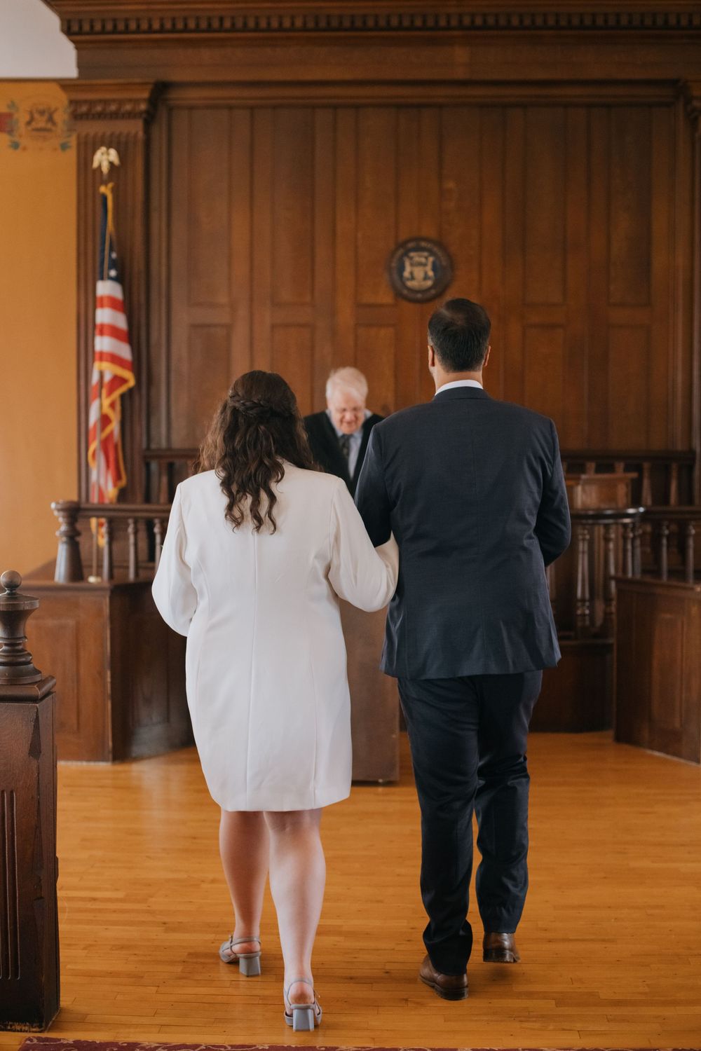Couple walks down courthouse aisle towards judge during civil wedding ceremony with American flag visible.