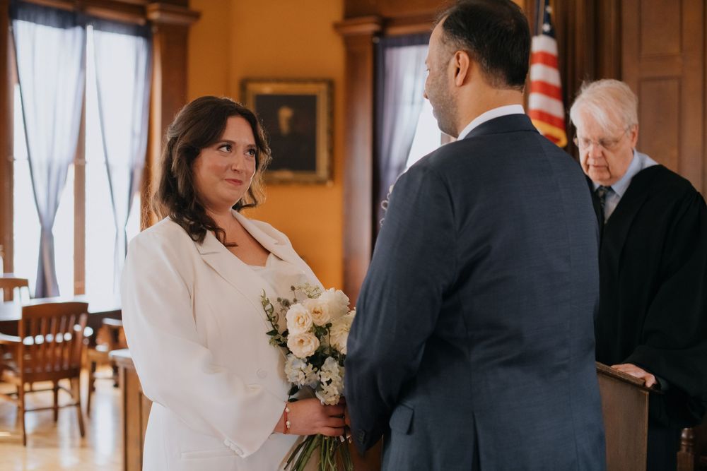 A couple stands facing each other during an indoor wedding ceremony with wooden chairs and an American flag visible.