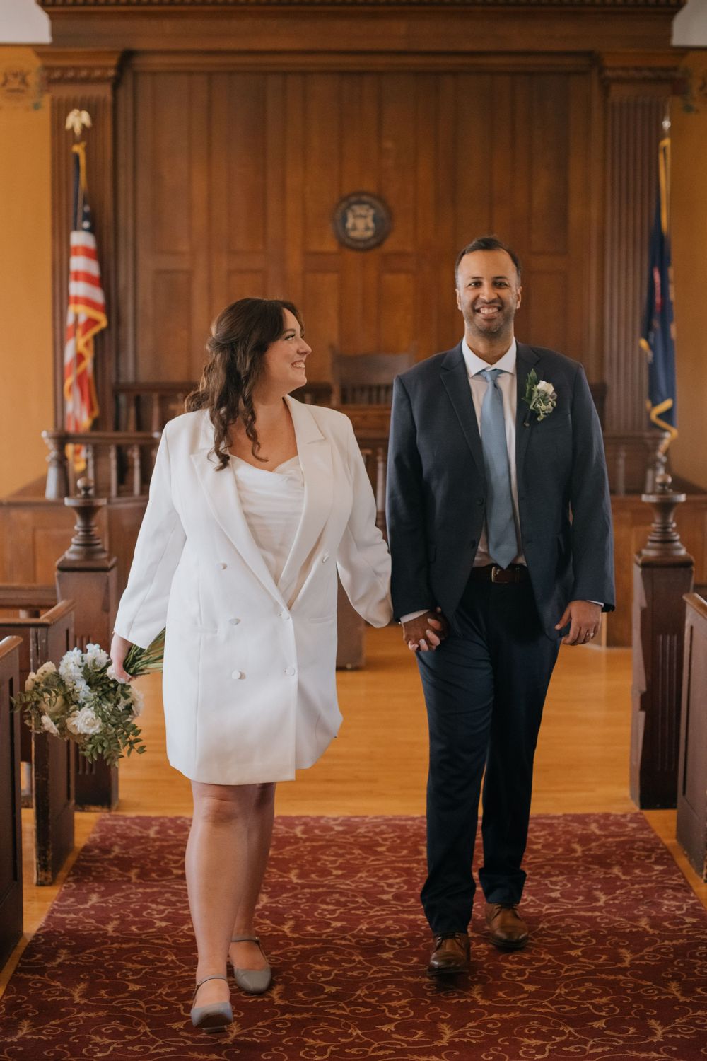 A romantic couple laughs and walks together across a carpeted hall with wooden paneling and an American flag.