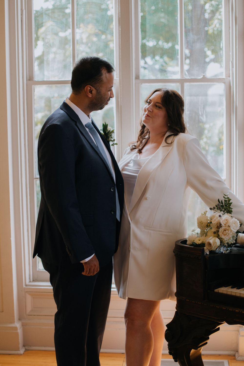 Couple shares intimate moment by window at indoor wedding venue with bouquet of white flowers on table.