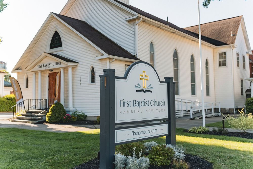 Exterior view of a white First Baptist Church building with a sign post in the foreground and green grass landscaping.