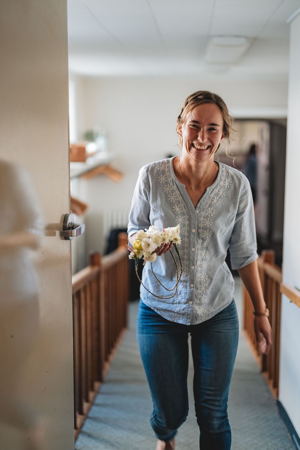 A person in a casual chambray shirt and jeans walks down a hallway holding white flowers with a joyful expression.