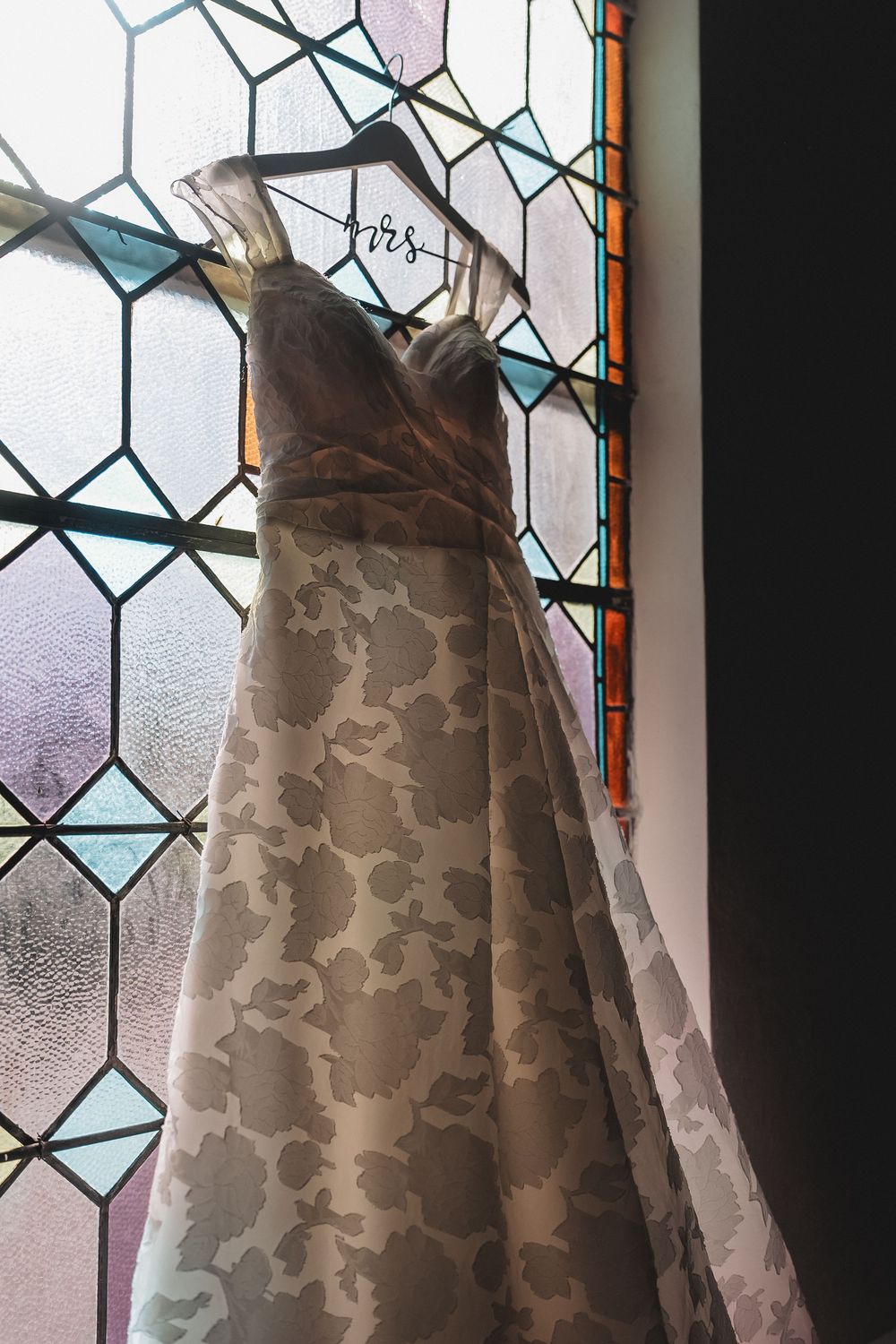A white wedding dress with floral lace pattern hangs elegantly against a stained glass window with hexagonal panes.