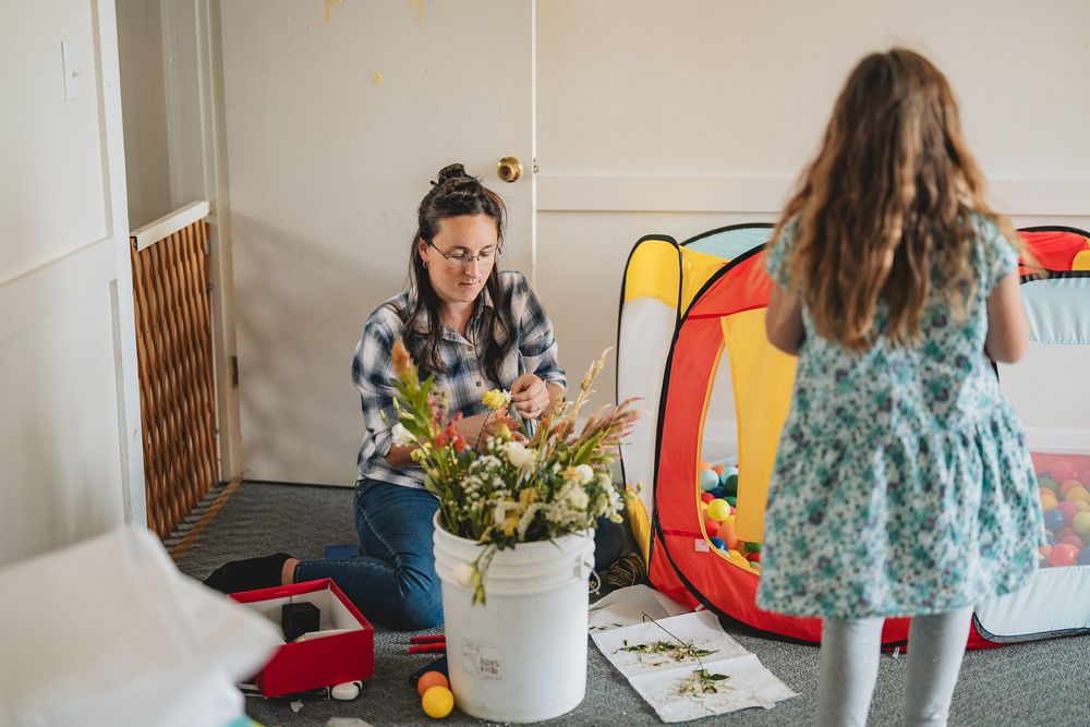 Two people arrange colorful flowers in a bucket while sitting in a room with toys and a portable play tent.
