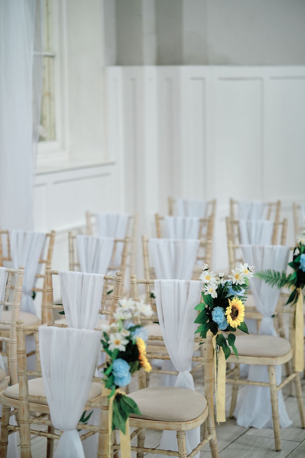 Elegant wedding ceremony setup with white chiffon chair drapes and greenery accents on gold chiavari chairs.