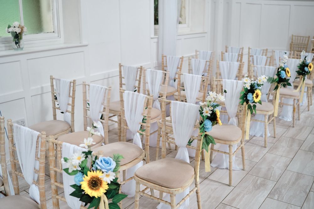 Light wood chiavari chairs with fabric sashes and sunflower accents line up in rows for an elegant indoor wedding ceremony.