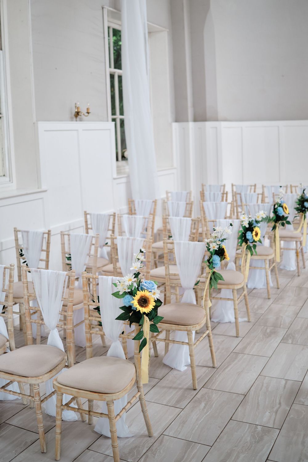 Elegant wedding ceremony setup with white fabric draped wooden chairs adorned with sunflower bouquets along the aisle.