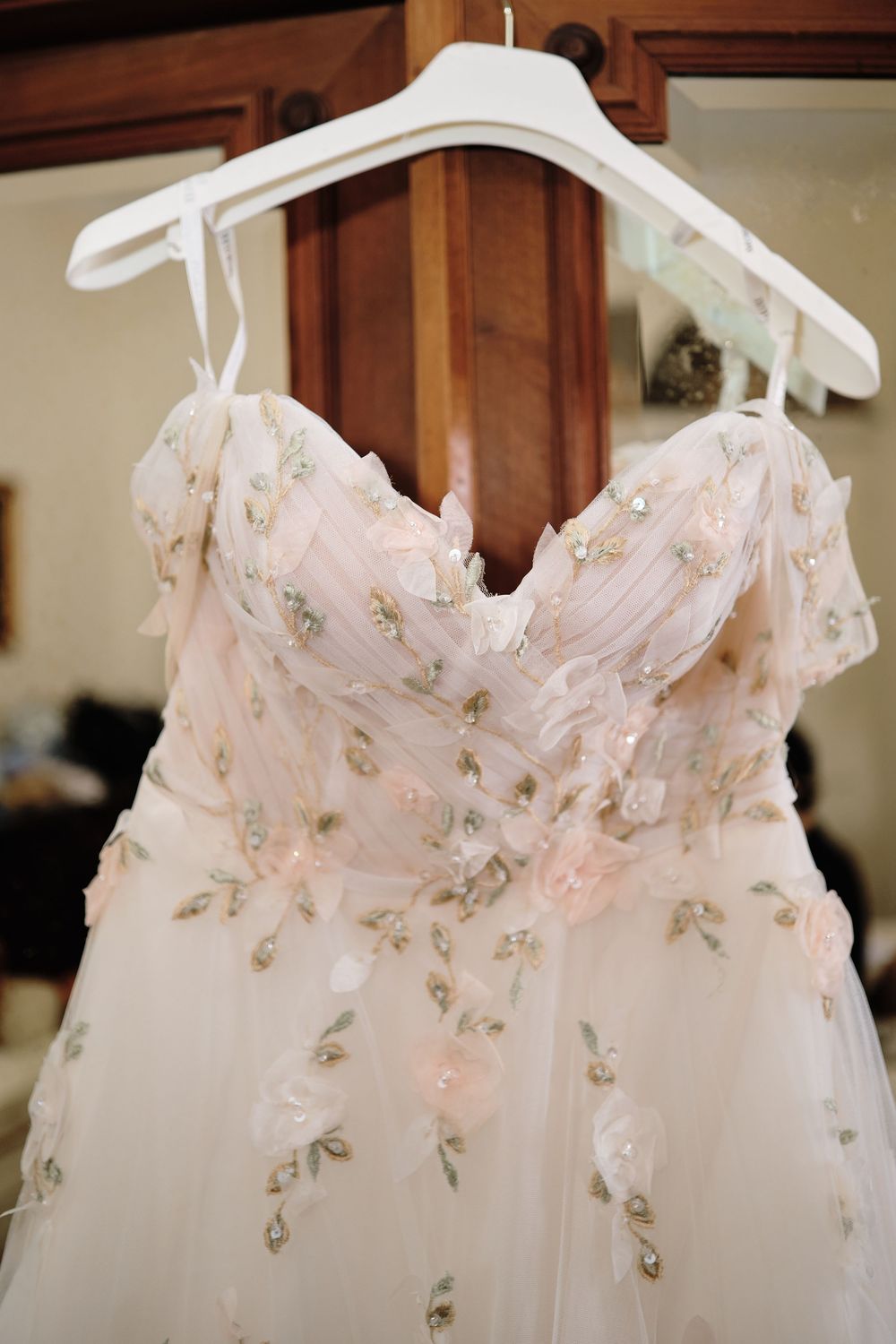 Delicate white wedding dress with floral embroidery details shown hanging on multiple hangers against wooden backdrop.