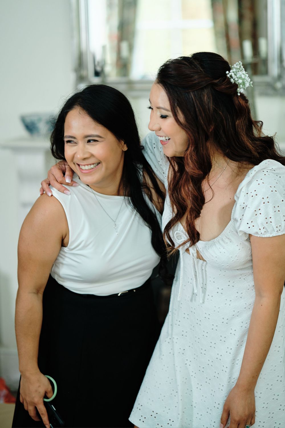 Two women in white outfits share a candid, joyful moment together in a bright indoor setting.