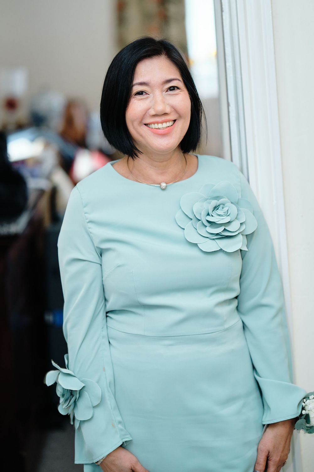Smiling woman wearing a mint green dress with floral accents and bell sleeves stands beside a white wall.