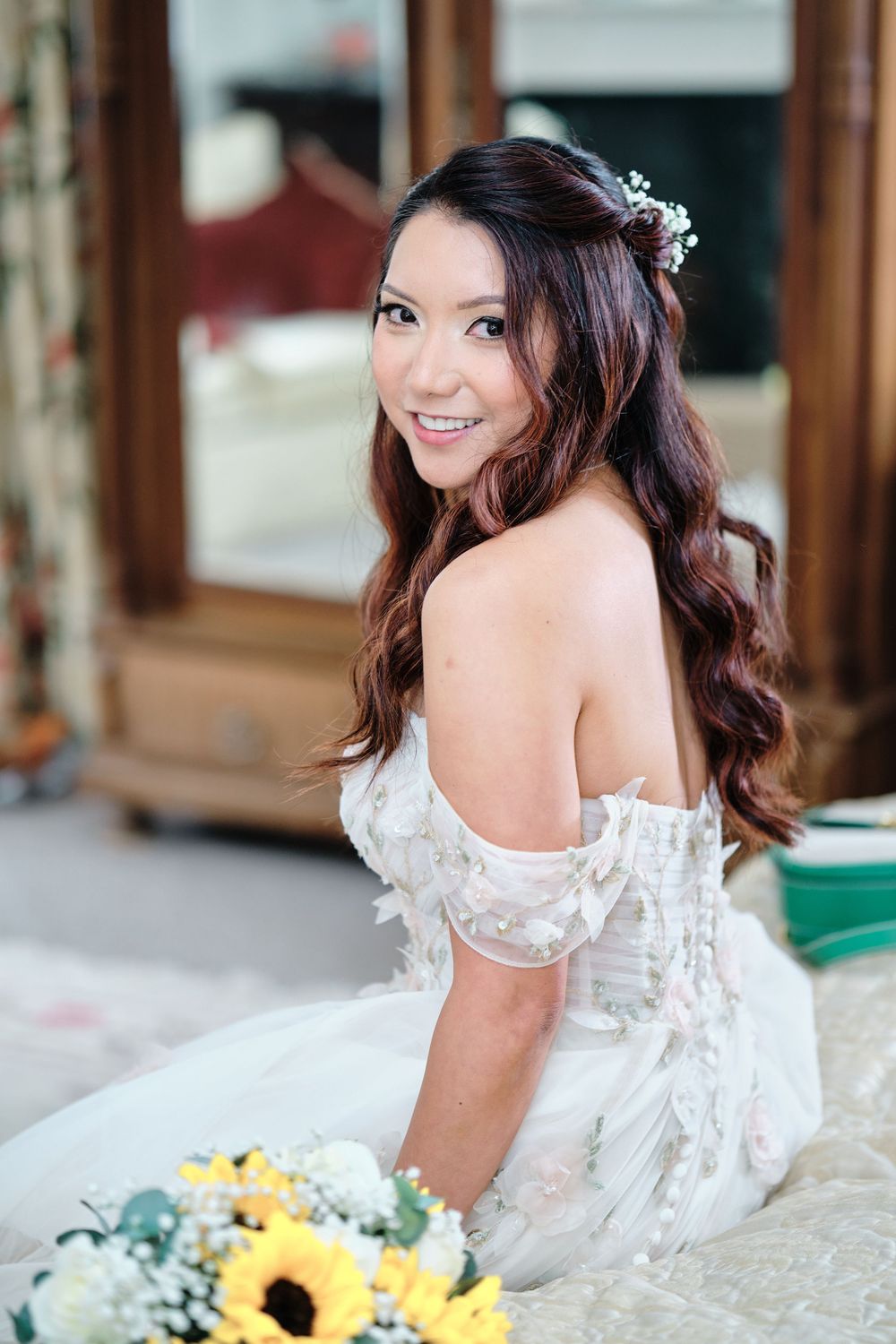 Beautiful bride in off-shoulder lace dress poses with sunflower bouquet in elegant indoor setting.