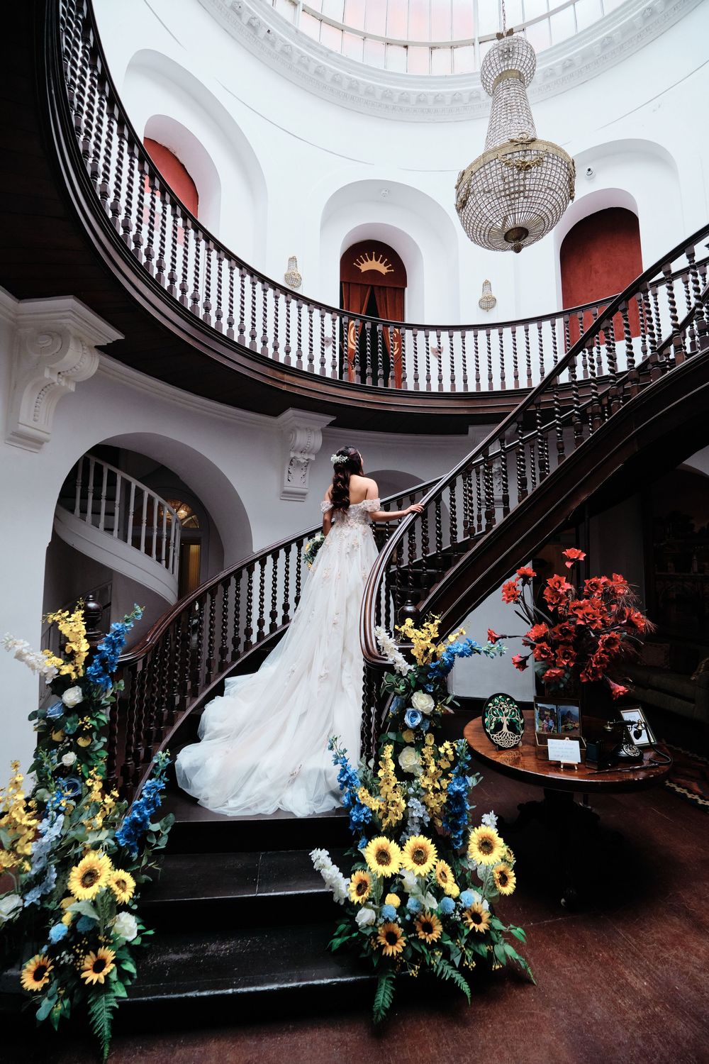 A bride in a flowing white gown poses on an elegant dark wooden staircase with sunflowers and blue floral arrangements.