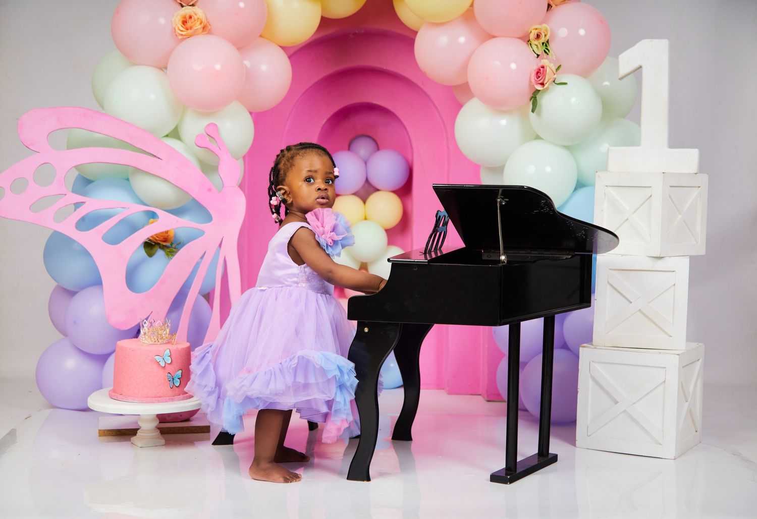 A young child in a purple dress plays at a grand piano surrounded by pastel balloon decorations.