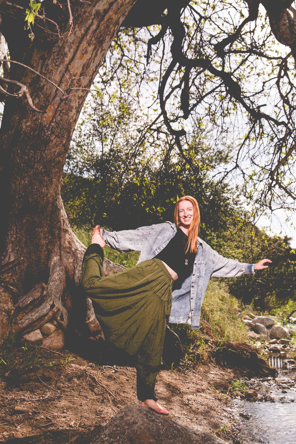 Woman in green skirt and denim jacket poses playfully by a large tree trunk near a stream in a natural setting.