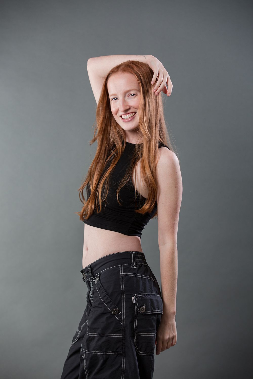 A person in a black crop top and dark cargo pants poses against a gray background with their arm raised.