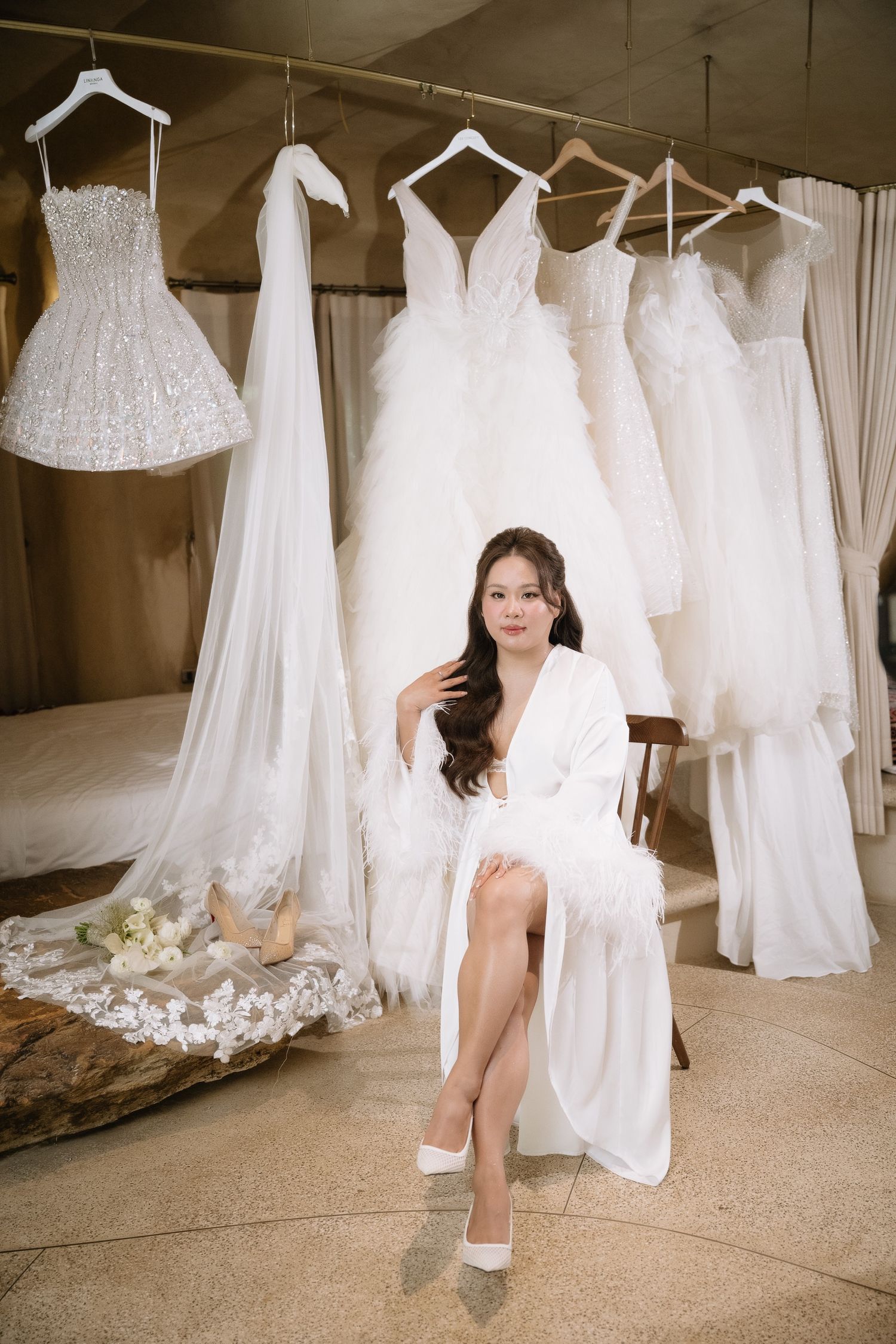 Bride in white feather robe sits elegantly surrounded by hanging wedding gowns in a bridal boutique.