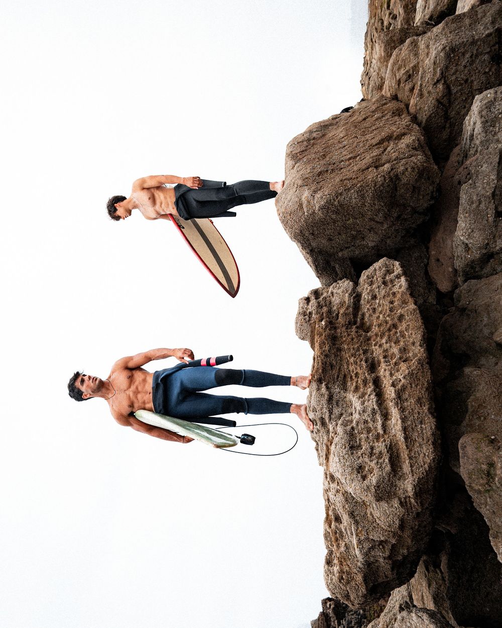 Two surfers with surfboards stand on rocky cliff formations against white background.