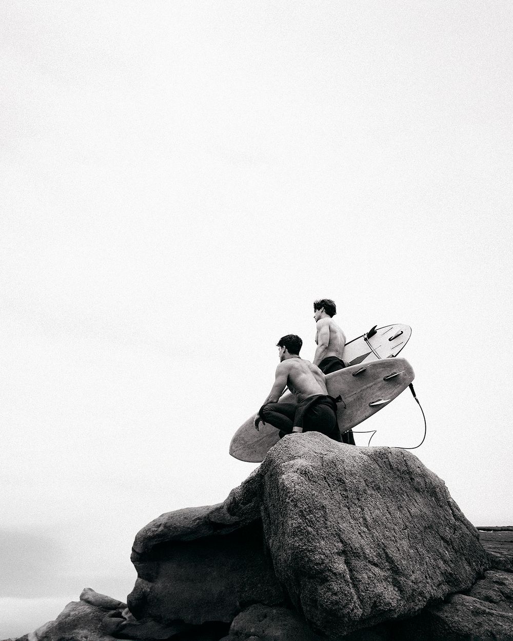 A person sits atop a large boulder with a surfboard while gazing at the ocean in a black and white photograph.
