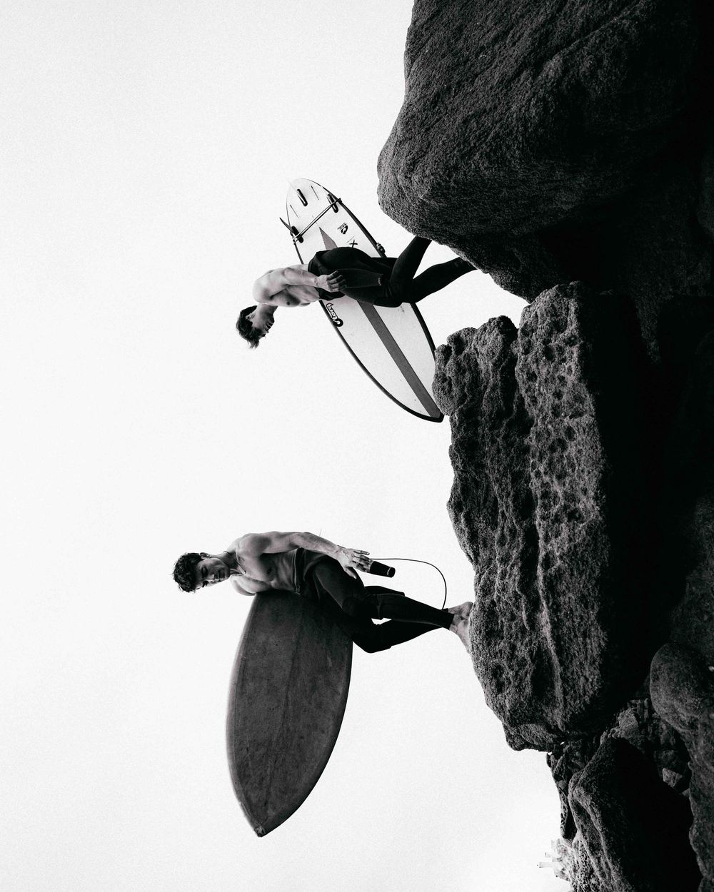 Black and white photo of surfers with boards climbing on dramatic coastal rocks.