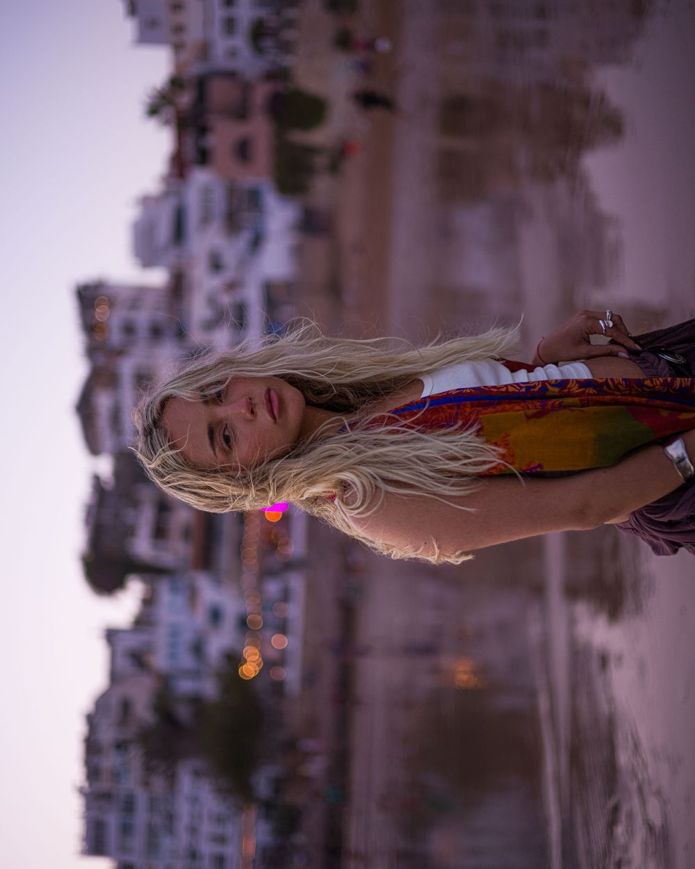 A person in colorful attire stands against a blurred cityscape background at dusk.