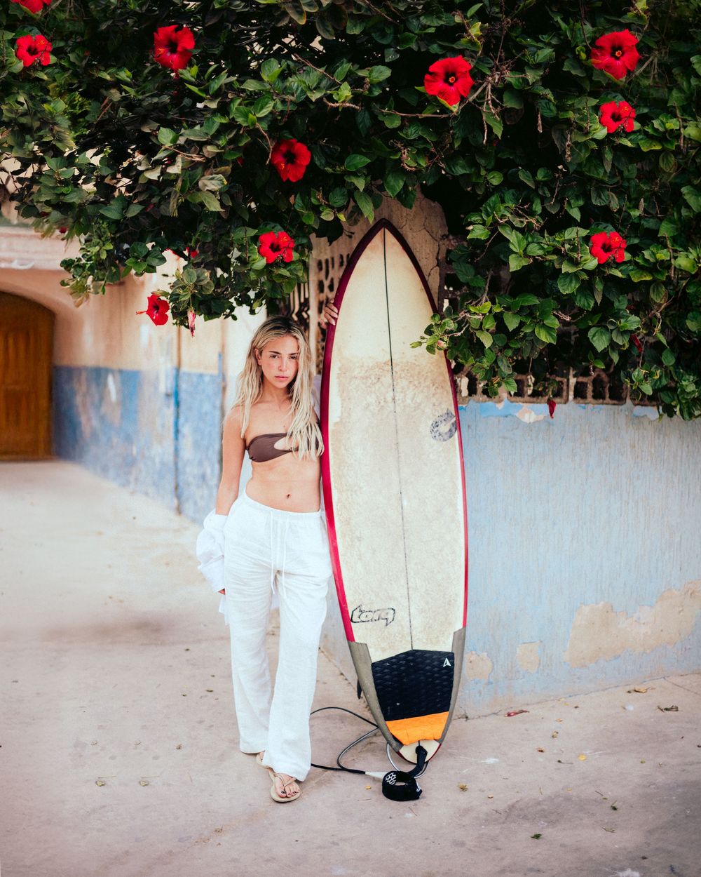 A series of surfboards leaning against a blue wall under red hibiscus flowers creates a tropical beach scene.