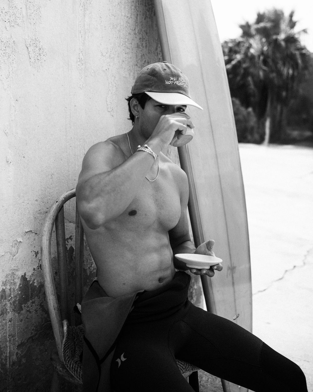 Shirtless surfer in a visor takes a coffee break while leaning against a wall with a surfboard nearby.