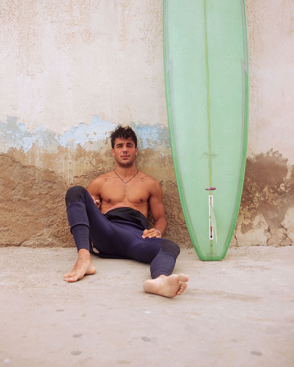 A surfer rests on sandy beach next to mint green surfboard against weathered wall.