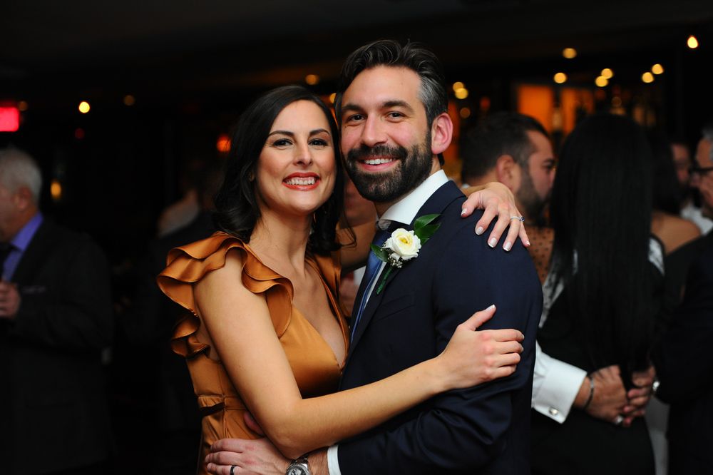 A couple in formal attire embrace and smile while dancing at an evening celebration with ambient lighting.
