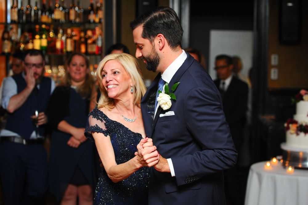Two people share a dance moment at a formal event, with a bar and bottles visible in the background.