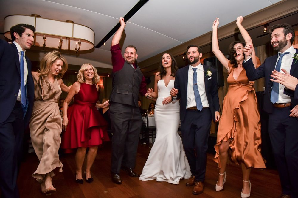 Wedding reception guests dancing energetically in a line with arms raised at an indoor venue.