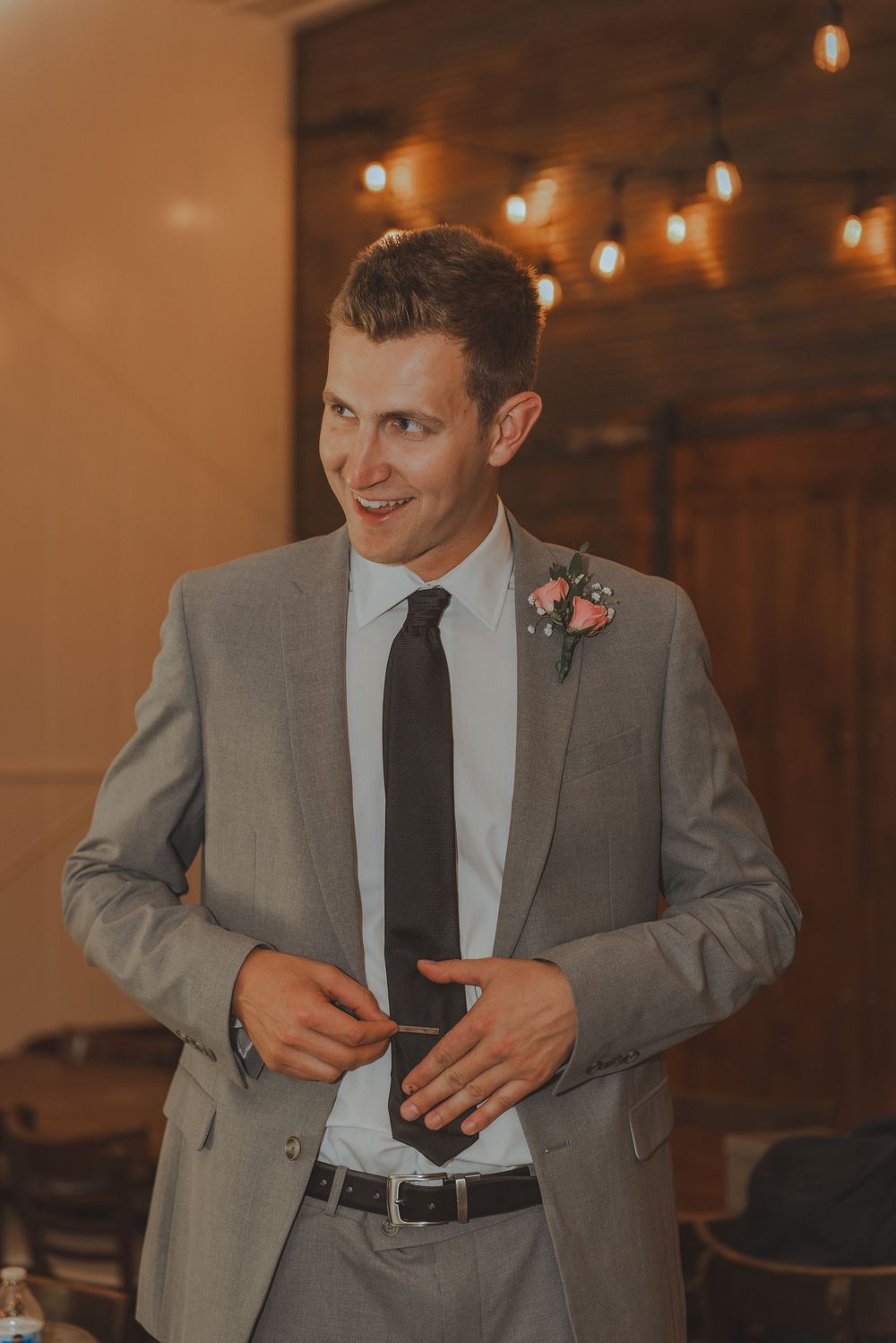 A groom in a grey suit and black tie smiles during an indoor wedding reception.