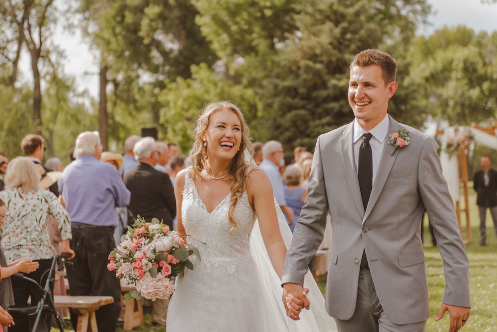 A joyful wedding couple walks hand in hand surrounded by guests on a sunny day.