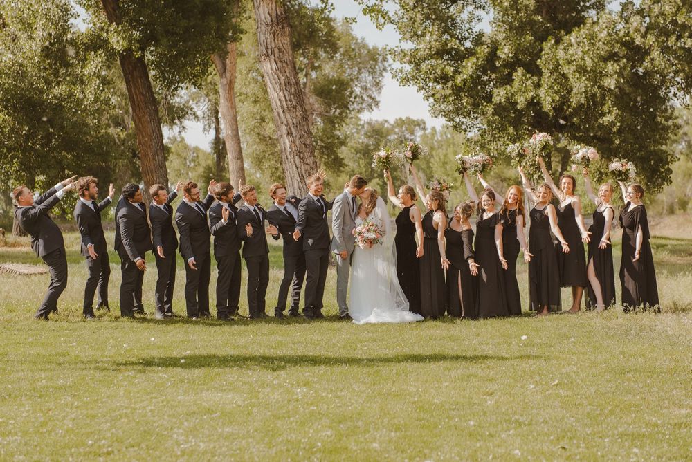 A large wedding party lined up for photos outdoors under tall trees.