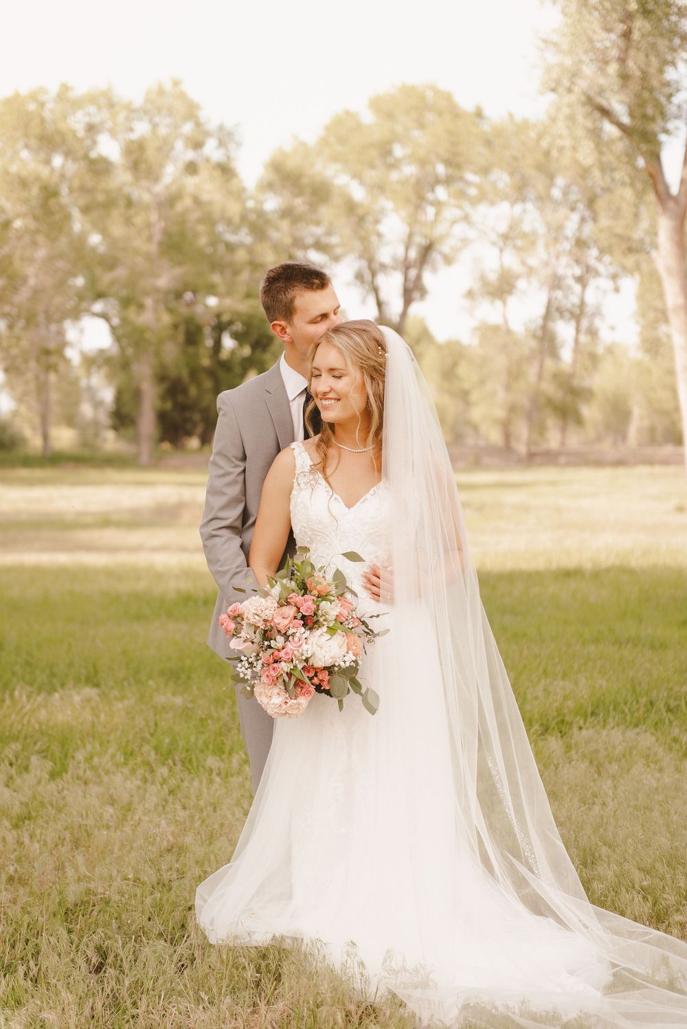 Newlywed couple shares intimate moments in a grassy field with pink floral bouquet and long veil.