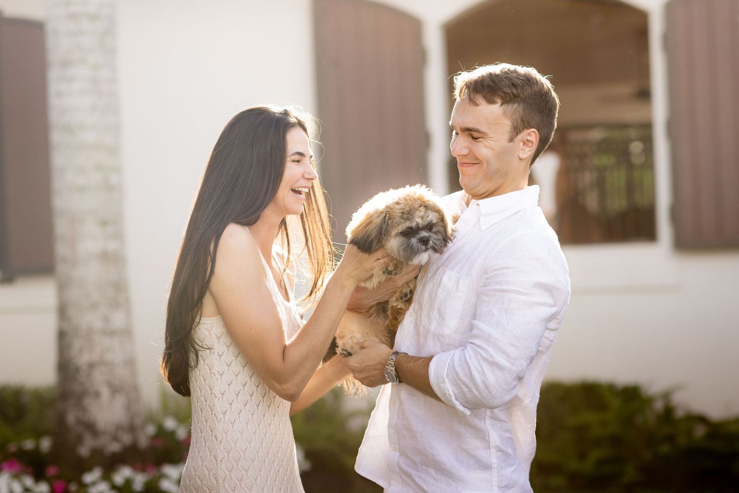 Couple playing with small dog outside white brick building in warm sunlight.