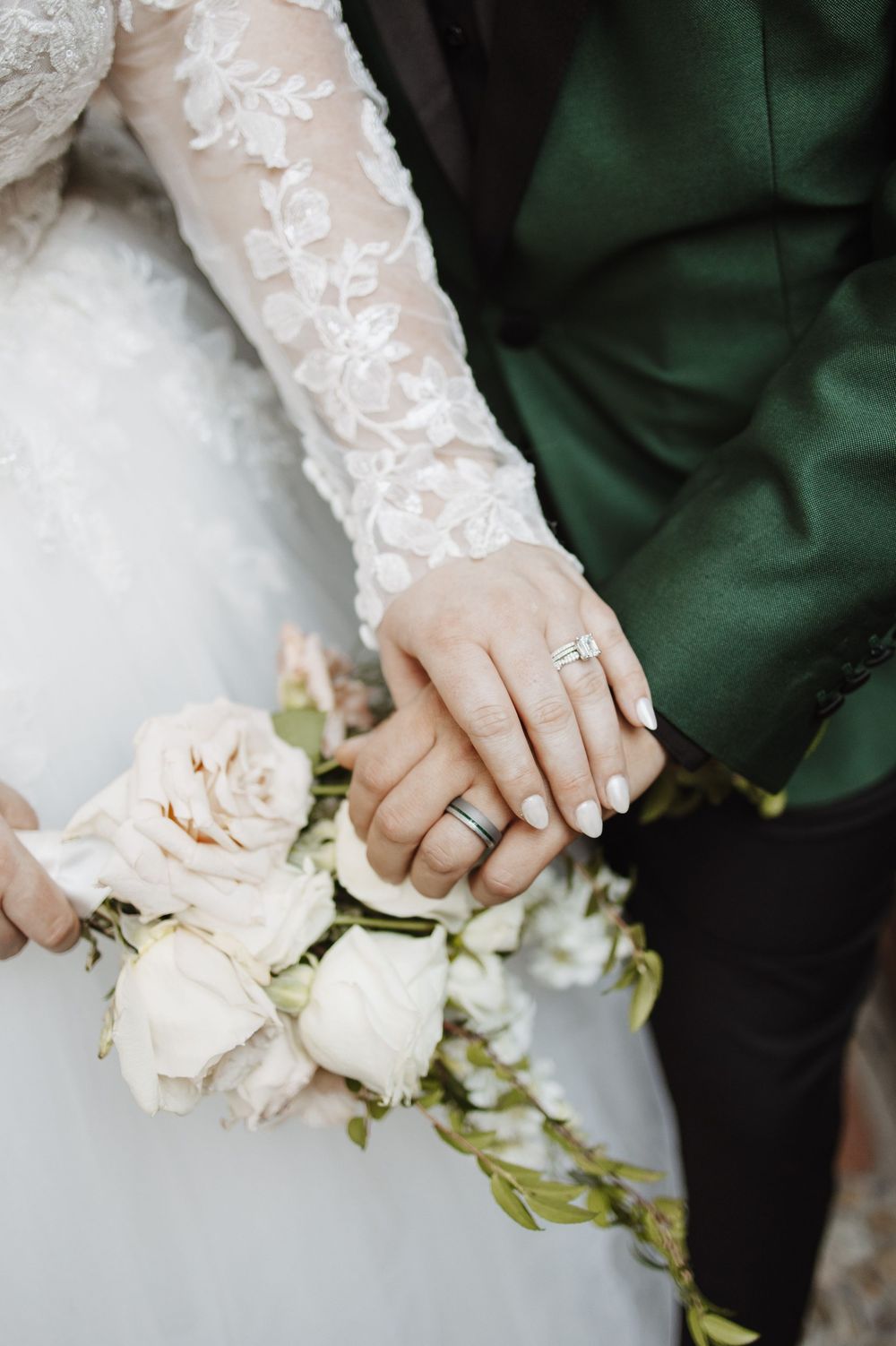 Close-up of bride and groom holding hands showing lace sleeve detail, engagement ring, and white rose bouquet.