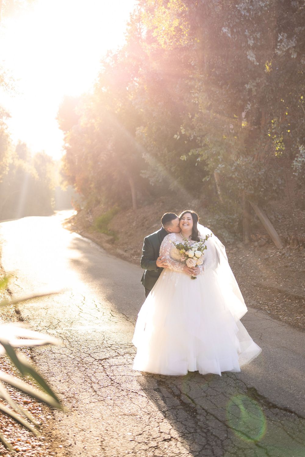 Wedding couple embraces in glowing sunlight on forest path during golden hour photo session.