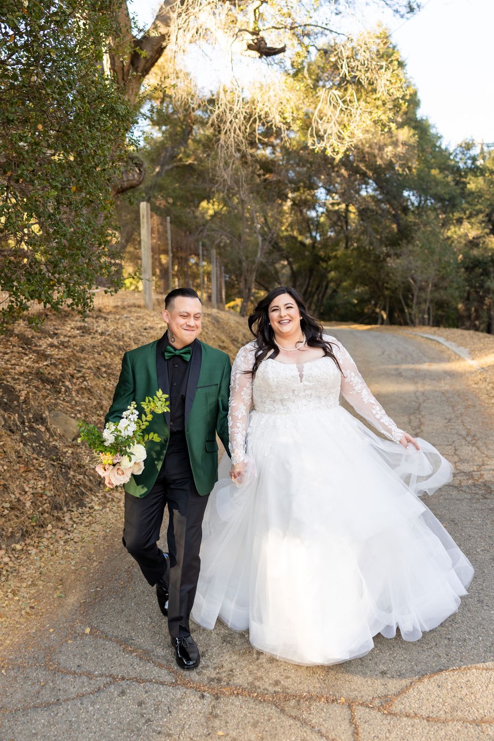 A couple in wedding attire walks along a dirt path surrounded by autumn trees as sunlight filters through the branches.