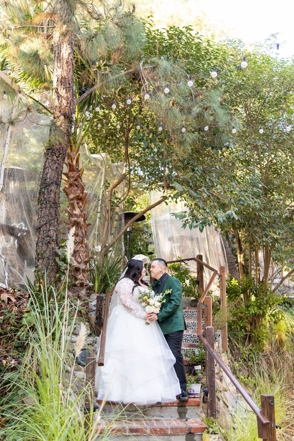A couple in formal wedding attire share an intimate moment in a lush garden setting surrounded by greenery and trees.
