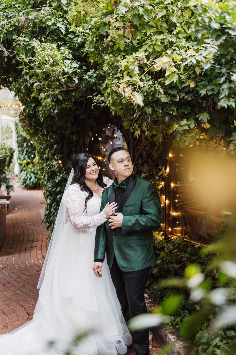 Couple shares an intimate moment under greenery on a brick patio with romantic string lights glowing in the background.