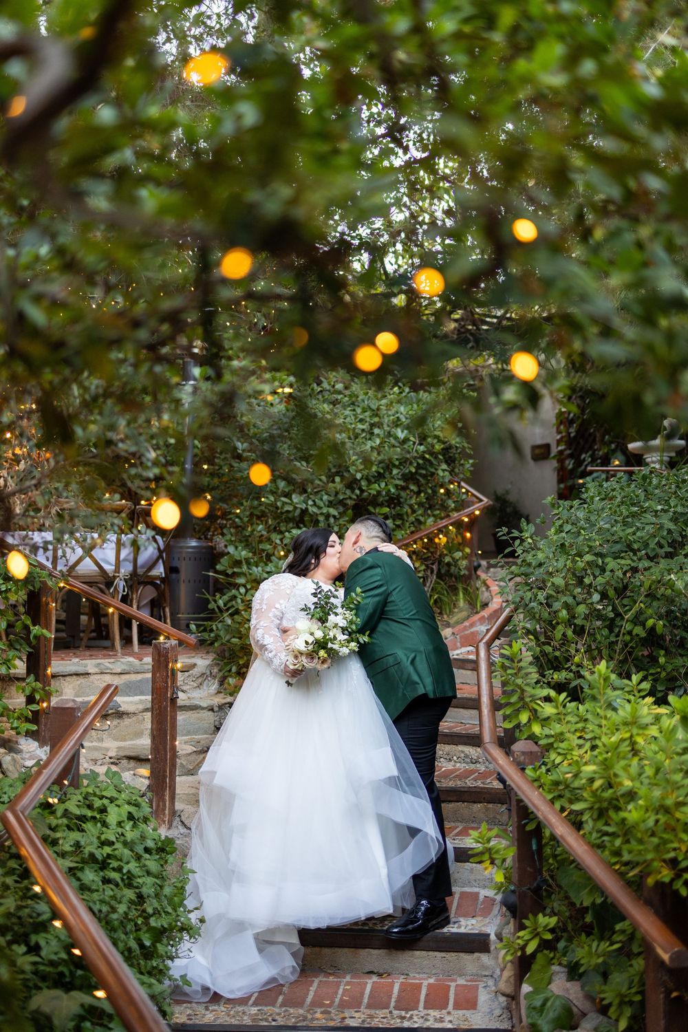 A couple in formal wedding attire shares a romantic moment under twinkling lights at dusk in a garden setting.