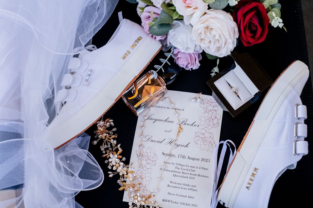 Wedding accessories flat lay with invitation card, white sneakers, tulle veil, and pink and red roses on dark surface.