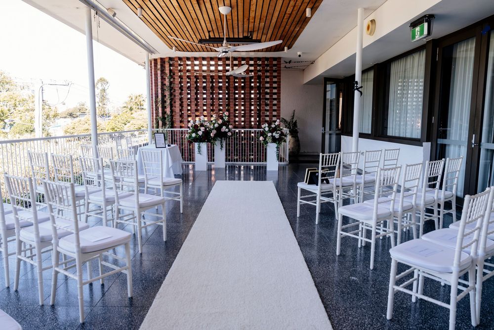 White chairs line both sides of a white aisle runner on an outdoor terrace set up for a wedding ceremony.