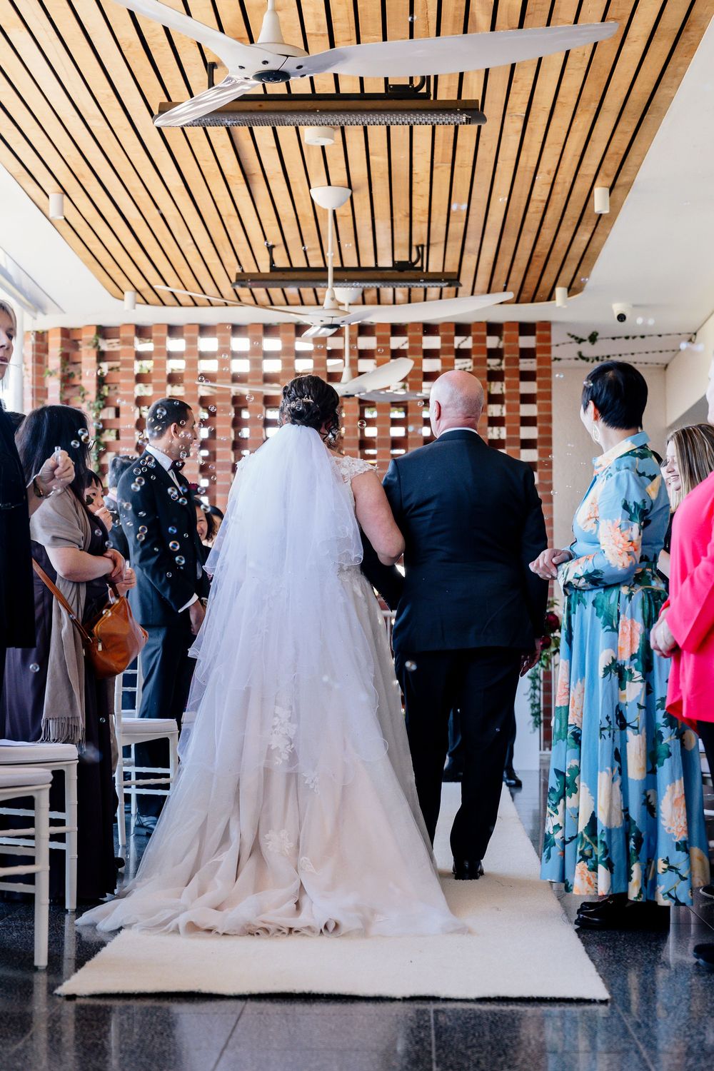 Wedding reception guests gather in a modern venue with wooden slat ceiling and white chairs.