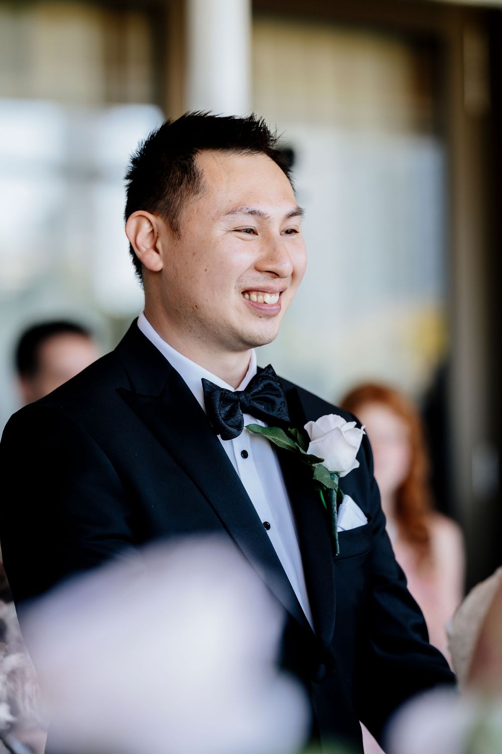 A groom at a wedding celebration laughs while wearing a black tuxedo with a white boutonniere during a formal reception.