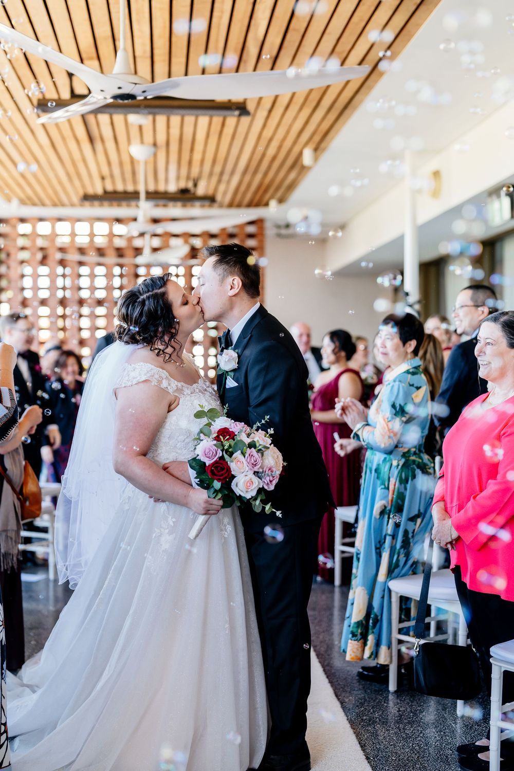 Wedding couple and guests celebrate during reception in modern venue with wooden slat ceiling and natural lighting.