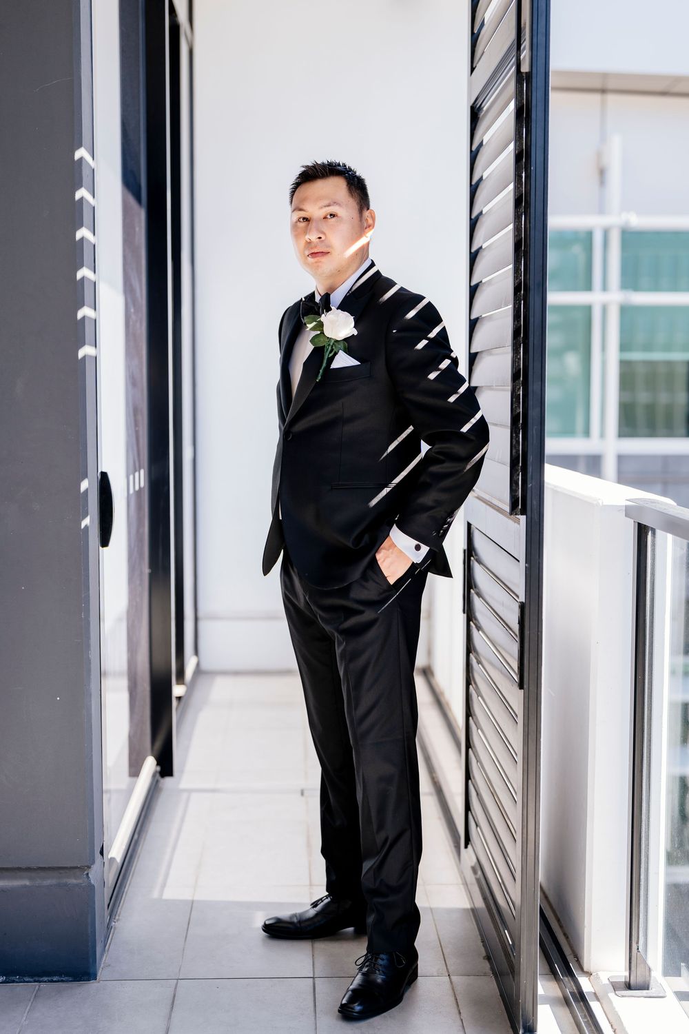A groom poses in a sleek black tuxedo in a bright modern hallway during formal wedding photography portraits.