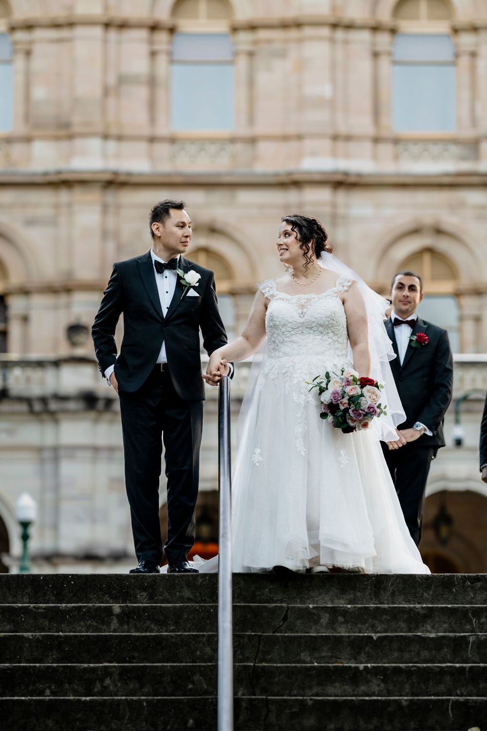 A joyful wedding couple descends stone steps at a grand historic building with arched architecture in the background.