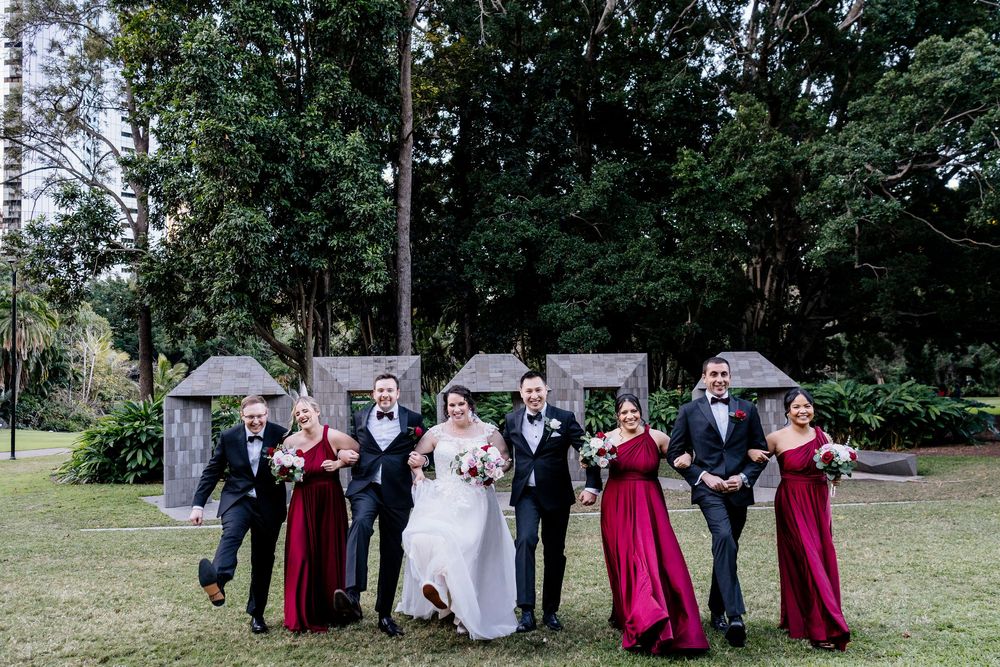 Wedding party poses together on grass with trees in background wearing dark suits and burgundy dresses.
