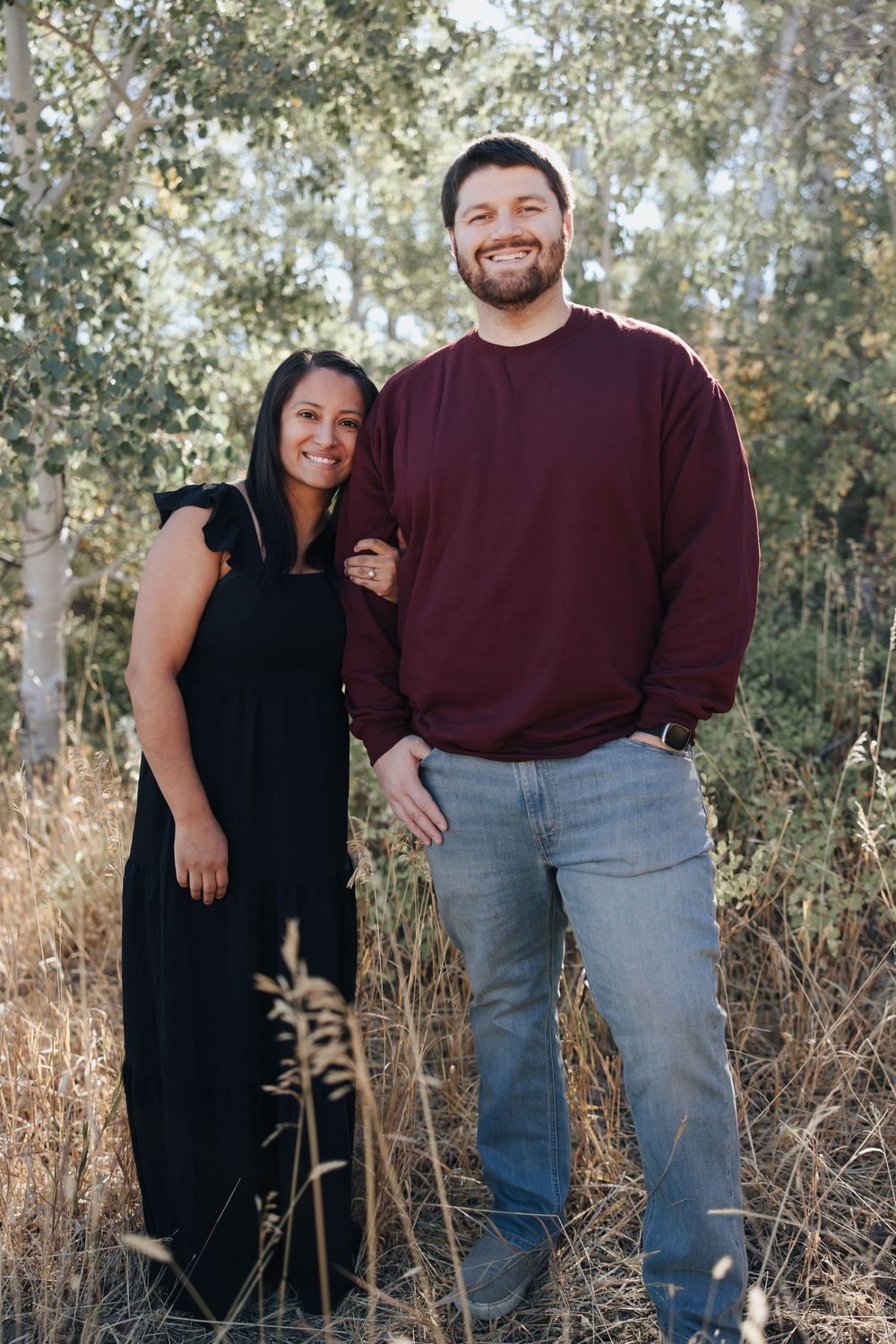 A couple poses together outdoors among tall grass and trees wearing burgundy and black clothing.